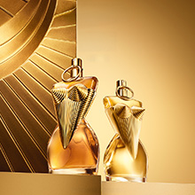 Zwei goldfarbene Parfumflakons von Jean Paul Gaultier in Form eines weiblichen Torsos, verziert mit auffälligen, strukturierten goldenen Brüstelementen. Die Flakons stehen vor einem luxuriösen, goldenen Hintergrund mit Sonnenstrahlen-Muster.