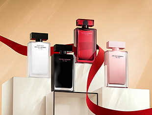 Vier Parfumflakons der For Her-Kollektion von Narciso Rodriguez in Rosa-, Rot-, Schwarz- und Weißtönen. Die Flakons stehen auf gestaffelten Podesten, umrahmt von einem geschwungenen roten Band vor warmem beigem Hintergrund.